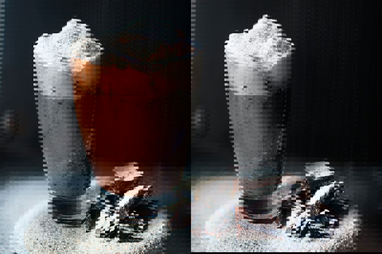 Et glas varm chokolade med skum og kakaodrys ved siden af chokoladestykker dækket af flormelis.