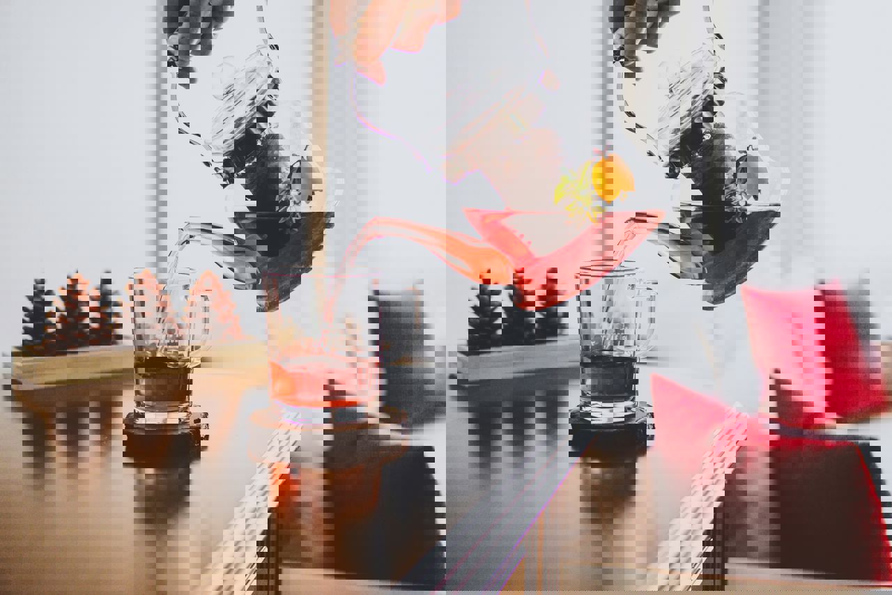 En person hælder rød frugtte fra en glaskande ned i en glaskop på et bord, med pyntekogler og puder i baggrunden.