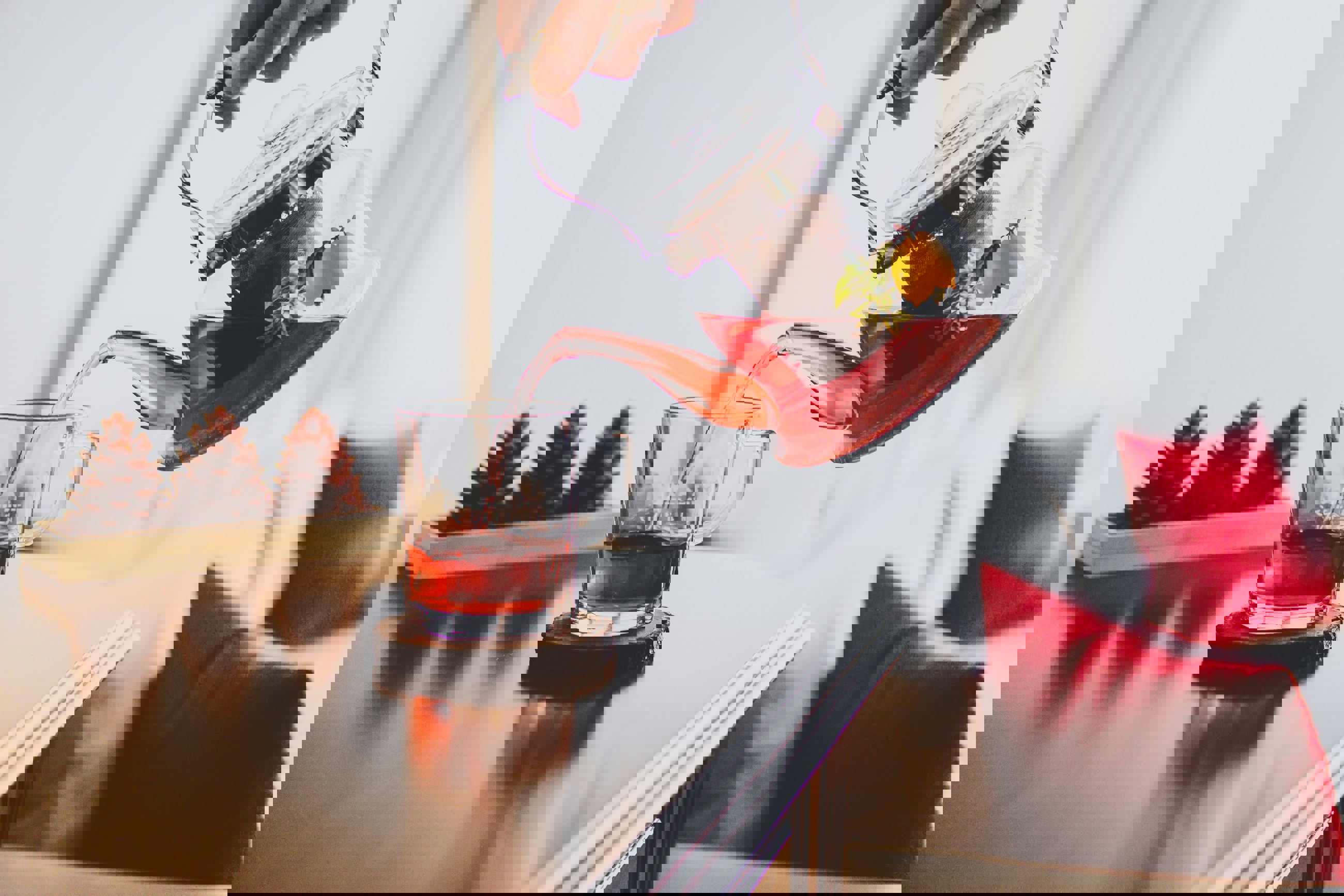 En person hælder rød frugtte fra en glaskande ned i en glaskop på et bord, med pyntekogler og puder i baggrunden.