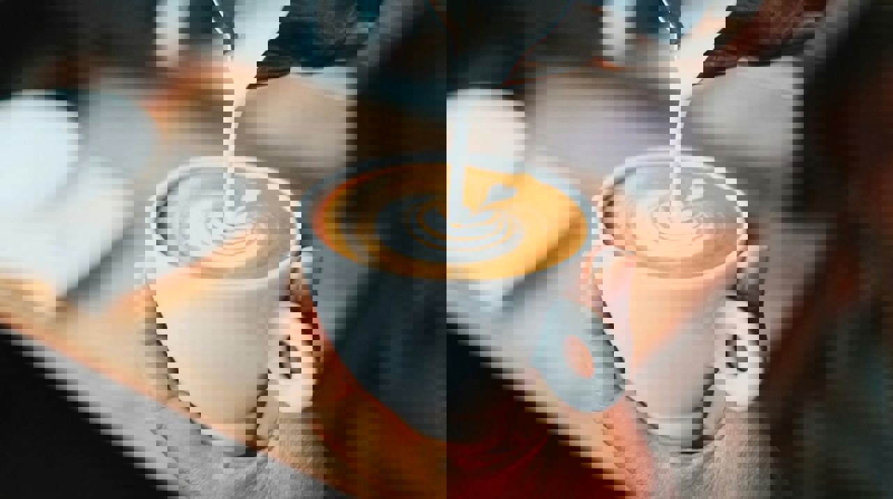 Nærbillede af en hånd, der holder en hvid kop cappuccino, mens mælk hældes i og danner latte art.