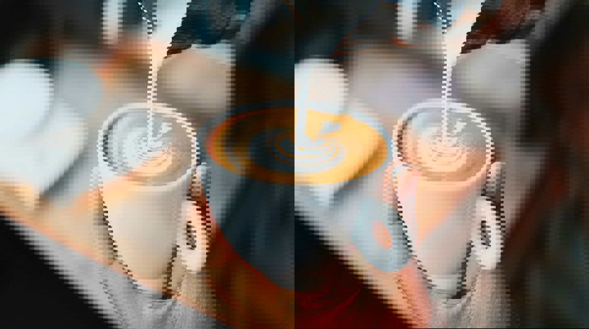 Nærbillede af en hånd, der holder en hvid kop cappuccino, mens mælk hældes i og danner latte art.